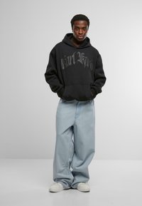 Sudadera negra con texto gráfico en el frente, combinada con jeans de pierna ancha de color azul claro. El modelo está de pie con las manos en los bolsillos, usando zapatillas blancas.