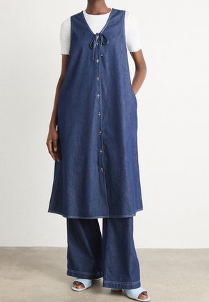 Person iført en ærmeløs knapfront denimkjole over flarede denim bukser og hvid kortærmet top med lyseblå åbne tå hæle.