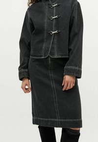 Frau in schwarzer Jeansjacke mit drei silberfarbenen Hakenverschlüssen und passendem schwarzen Jeansrock, steht mit entspannten Händen.