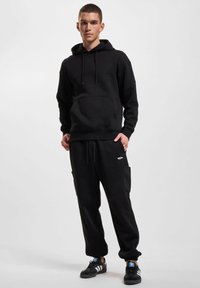 Svart hettegenser med teksturert øvre del, snorhette og kengurulomme, kombinert med sorte joggebukser med elastisk liv som har sidelommer.