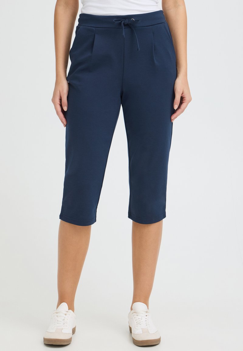 Femme portant un pantalon à cordon de serrage bleu marine mi-genoux et des baskets blanches, debout devant un fond clair uni.
