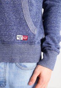 Nahaufnahme einer Person, die ein blaues Superdry Track & Field Sweatshirt und hellblaue Jeans trägt, mit einer Hand, die neben der Tasche ruht.