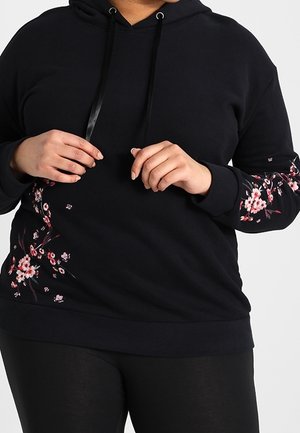 Sort hættetrøje med pink blomsterprint på ærmer og side, båret af en person, der justerer snoren.