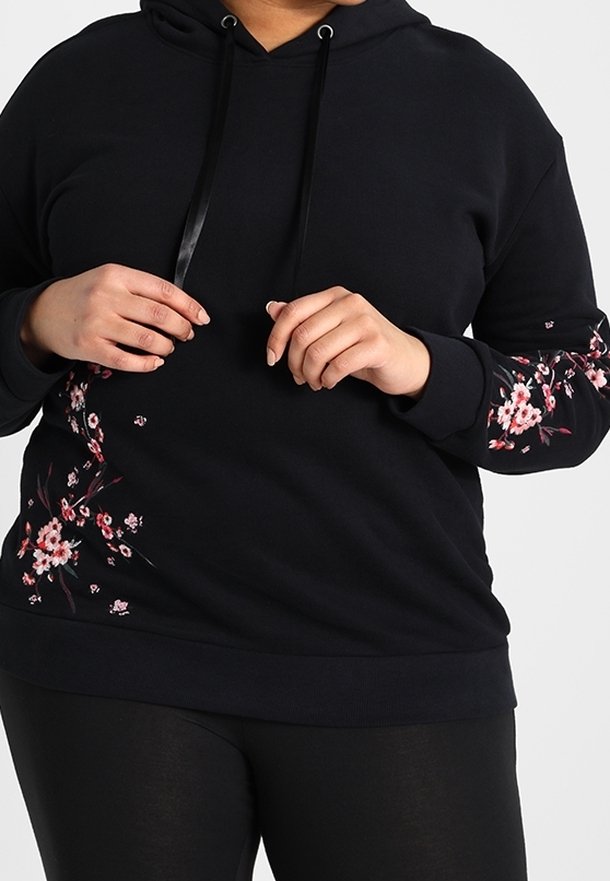 Sweat à capuche noir avec motifs floraux roses sur les manches et le côté, porté par une personne ajustant le cordon.