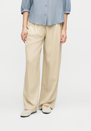 Donna che indossa pantaloni beige a gamba larga, camicia azzurra chiara a maniche lunghe con bottoni, e scarpe basse di colore chiaro, in piedi davanti a uno sfondo semplice.