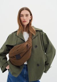 Borsa a tracolla marrone in morbido tessuto, con una consistenza sottile e una patch con logo nera. Il modello indossa una giacca verde oversize.