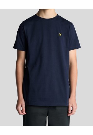 Personne portant un t-shirt bleu foncé uni à manches courtes avec un petit logo d'oiseau jaune sur la poitrine gauche, debout devant un fond uni.