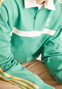 Grön zip-up sweatshirt med vita och gula ränder, med en krage och Adidas-logotyp på bröstet, tillverkad av ett mjukt, strukturerat tyg.