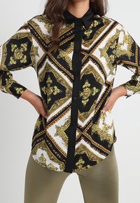 Košile s dlouhým rukávem zdobená výrazným barokním vzorem v černé, bílé a zlaté barvě, s zapínáním na knoflíky vpředu a černým límečkem. Hladká látka, pohodlný střih.