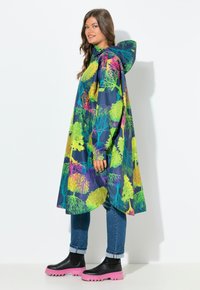 Vrouw in een gewatteerde regenjas met kleurrijke boompatronen, blauwe jeans en zwarte laarzen met dikke roze zolen, staande in de zij-aan-zij houding.