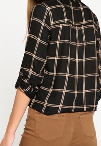 Žena oblečená v černém a béžovém kostkovaném dlouhém rukávu s vyhrnutými rukávy a hnědých kalhotách, viděná zezadu.
