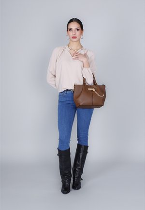 Borsa a tracolla in pelle marrone con dettaglio a catena, abbinata a una blusa beige chiara, jeans blu e stivali neri fino al ginocchio, ambientata su uno sfondo grigio.