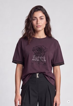 Woman with long wavy hair wearing a dark purple T-shirt with a rose design and black high-waisted pants against a plain light background.