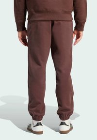 Pantaloni della tuta marroni con polsini elasticizzati e una vestibilità comoda, realizzati in materiale morbido. Le scarpe presentano un design bianco e nero con un accento a righe.