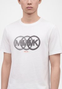 Άνδρας που φοράει λευκό T-shirt με μαύρες οριζόντιες ρίγες που επικαλύπτουν το λογότυπο MK και μικρό κόκκινο κείμενο Michael Kors από κάτω.