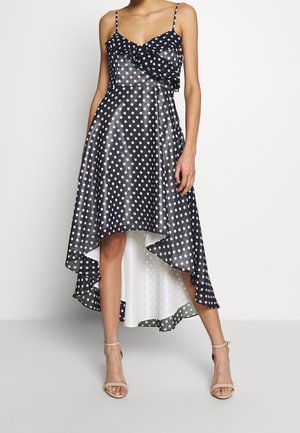 Femme portant une robe haute-bas à pois noir et blanc avec des bretelles spaghetti et des talons beige à bride chevillée, se tenant devant un fond uni.