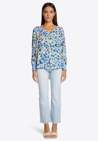 Bloemige blouse met een v-hals ontwerp, voornamelijk in blauw en geel, lange mouwen met blauwe manchetten, gecombineerd met lichtblauwe spijkerbroeken.
