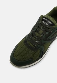 Sneaker in rete e pelle verde e nera con lacci neri, suola bianca e etichetta "Scotch & Soda" sulla linguetta imbottita.