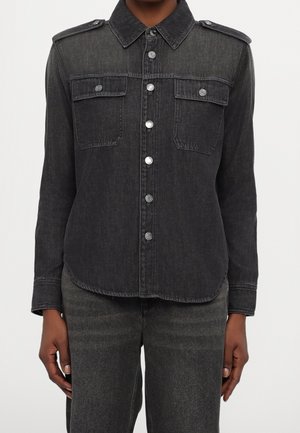 Person trägt ein kohlschwarzes Jeanshemd mit Knopfleiste, zwei Brusttaschen, Schulterklappen und passende dunkle Jeans.