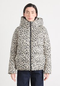 Eine Person steht in einer gepolsterten Jacke mit Leopardenmuster und großer Kapuze sowie dunklen Jeans, posierend vor einem hellen Hintergrund.