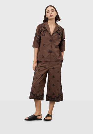 Žena oblečená v hnedej košeli na gombíky a v zladených širokých skrátených nohaviciach s čiernymi vyšívanými kvetinovými vzormi, stojaca s uvoľnenými rukami.