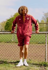 Burgunderfarget treningsdress med gule detaljer, som har en glidelåsjakke og shorts. Bæres med hvite joggesko, ved en kjettinggjerde.