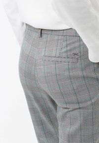 Grå rutiga byxor med ett rutig mönster som har tunna gröna linjer, rak passform och en bakficka. Tyget har en synlig struktur.