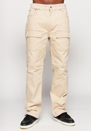 Pantalon cargo beige avec grandes poches avant à rabat, porté avec des baskets noires sur une personne debout sur un fond uni.