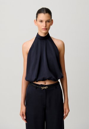 Donna che indossa un top nero senza maniche a collo halter e pantaloni neri a vita alta con una piccola cintura decorativa a fiocco, in piedi davanti a uno sfondo semplice.