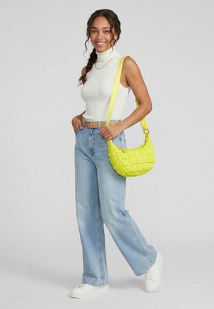 Woman wearing white sleeveless turtleneck, light blue jeans, white sneakers, carrying a bright yellow quilted shoulder bag, smiling.