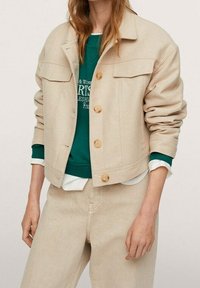 Veste courte beige avec deux poches poitrine, boutons en bois et manches retroussées, portée sur un sweat-shirt vert et un pantalon de couleur crème.