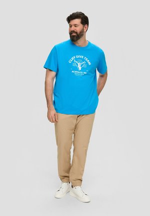 Blaues Baumwoll-T-Shirt mit dem Schriftzug "CLIFF DIVE TEAM", getragen mit beigen Hosen und weißen Sportschuhen. Das Shirt hat kurze Ärmel und einen Rundhalsausschnitt.
