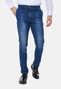 Jeans in denim blu slim-fit con una texture liscia, dettagli sbiaditi e chiusura con bottone, abbinati a scarpe eleganti nere lucide.