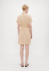 Short-sleeve dress in light beige with a shell-pattern print, cinched waist, and knee-length hem, made of soft fabric. Simple flat sandals.