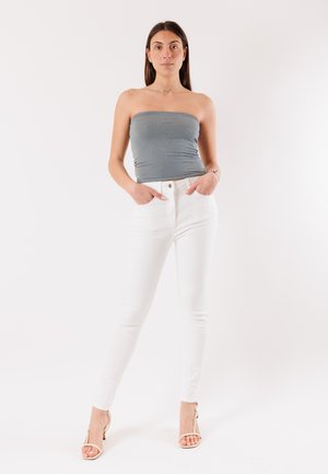 Donna in piedi con le mani in tasca, indossa un top grigio senza spalline, pantaloni bianchi e sandali beige con tacco alto su uno sfondo semplice.