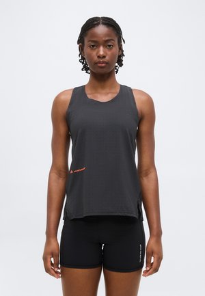 Jonge vrouw met gevlochten haar, gekleed in een zwarte mouwloze sporttop en zwarte short, staand met het gezicht naar voren tegen een effen achtergrond.