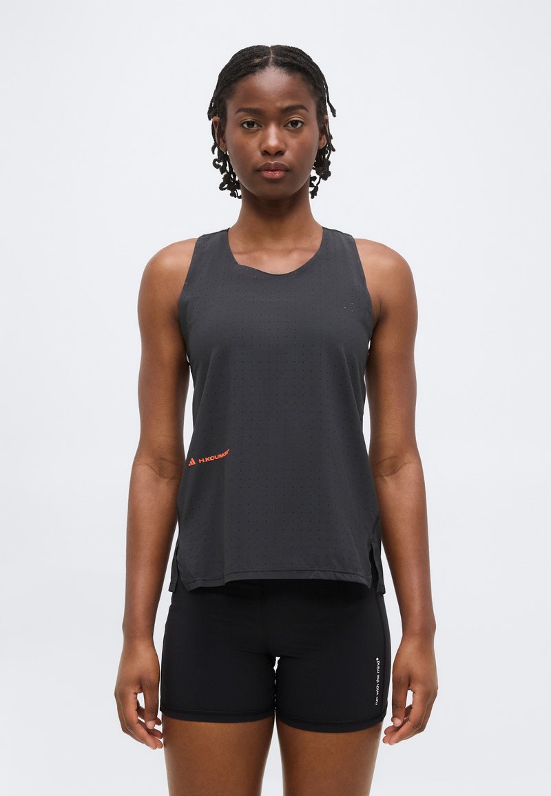 Jeune femme aux cheveux tressés portant un haut de sport noir sans manches et un short noir, debout face à un fond uni.