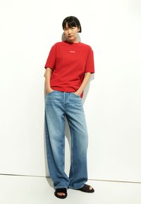 Rotes Baumwoll-T-Shirt mit kurzen Ärmeln und weißem Text. Hellblaue weite Jeans mit fünf Taschen. Schwarze Sandalen. Einfarbiger weißer Hintergrund.