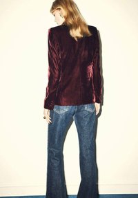 Blazer de terciopelo burdeos con hombros marcados, combinado con jeans de denim azul acampanados, que muestran una textura suave y detalles de costura prominentes.