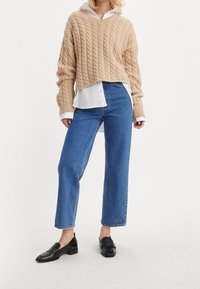 Suéter beige de punto con cuello en V y hombros caídos, combinado con jeans azul de pierna recta y mocasines negros. Camisa blanca visible debajo.