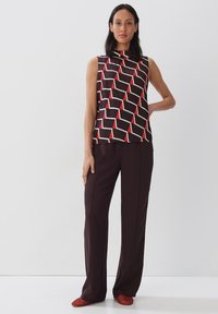 Mouwloos zwart top met een rood en wit geometrisch patroon, gecombineerd met donkerbruine broeken en rode schoenen. Glad materiaal met een modern ontwerp.