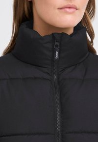 Svart polstret jakke med høy krage, glidelås og et merket glidelåsdrag. Stoffet ser glatt ut med en teksturert overflate.
