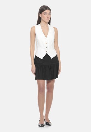 Donna che indossa un gilet bianco con bottoni sopra una gonna nera con orlo arricciato, abbinato a tacchi neri a punta, in piedi su sfondo bianco.