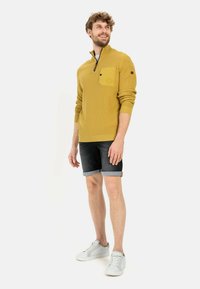 Senfgelber Pullover mit einem Reißverschlusskragen und Fronttasche, kombiniert mit dunklen Jeansshorts und weißen Sneakers. Leichte strukturierte Strickware.