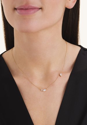 Delicado collar de cadena de oro con tres pequeñas gemas transparentes, llevado alrededor del cuello sobre ropa negra.