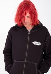 Sweat à capuche zippé noir avec des coutures blanches, présentant une poche avant et un logo circulaire "VOLCOM" sur le côté gauche de la poitrine.