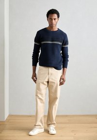 Marineblå strikkegenser med teksturert mønster og stripete detaljer, kombinert med beige bukser og hvite sneakers. Enkelt bakgrunn, tregulv.