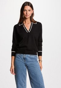 Donna che indossa un maglione nero a maniche lunghe con rifiniture bianche sul colletto e sui polsini, abbinato a jeans a vita alta di blu chiaro, in piedi dritta.