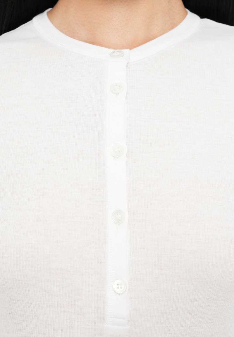 Maglietta bianca a maniche corte con colletto rotondo. Realizzata in tessuto testurizzato con sei bottoni bianchi lungo la chiusura.