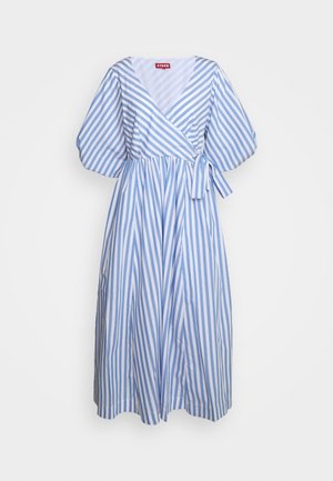 Nainen pukeutunut sinivalkoraidalliseen midimekkoon, jossa on liehuvat hihat, ja valkoisiin slingback-korkokenkiin, seisoo harmaalla lattialla seinää vasten.